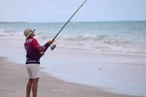 Angler wearing a wide-brim hat casts a fishing rod from a quiet sandy beach toward turquoise sea.
