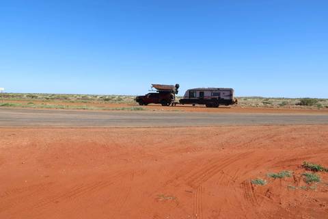 Pickup truck towing a caravan parked on a red-dust roadside in the Australian outback under a clear blue sky.