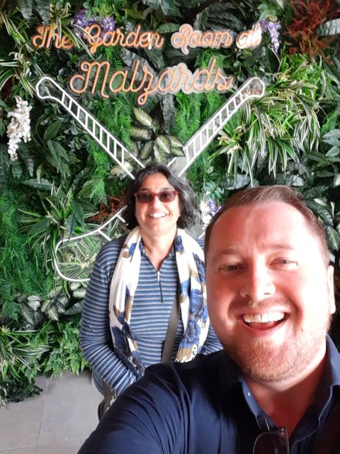 Close-up selfie of two smiling travellers against a lush indoor wall of green plants.