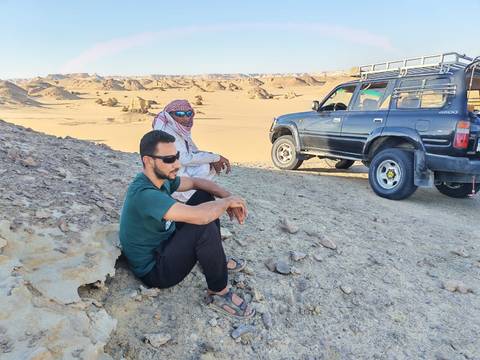 Two men rest beside an off-road vehicle amid Egypt’s sandy desert hills under bright sunlight.