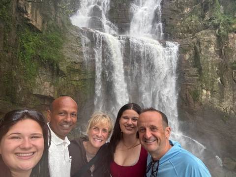 A smiling group poses for a selfie in front of a misty waterfall surrounded by rock walls.
