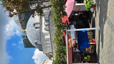 A grey–domed Ottoman-style mosque framed by small street food stalls and bright blue sky with scattered clouds.