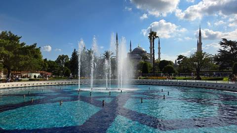 A circular fountain pool with high jets of water set before the silhouette of the Blue Mosque and minarets under a vivid blue sky.