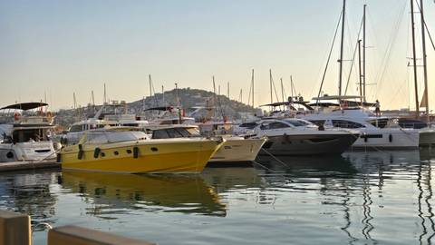 A calm marina filled with yachts and sailboats; a bright yellow motorboat stands out against distant hills.