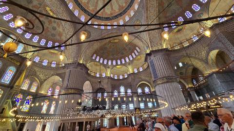 The ornate, domed interior of the Blue Mosque filled with worshippers and visitors under hanging lamps.