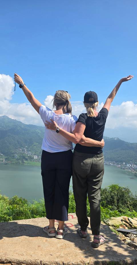 Two women embrace with arms raised atop a viewpoint overlooking a wide lake and green hills.