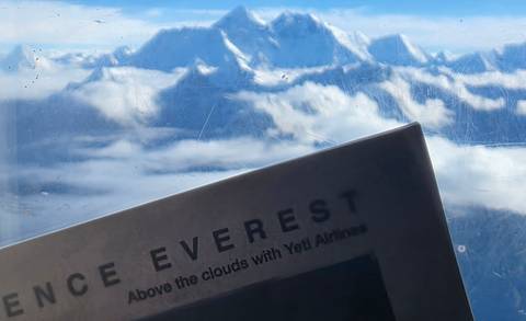View of snowy Himalayan peaks through a scratched plane window with an Everest flight brochure in the foreground.