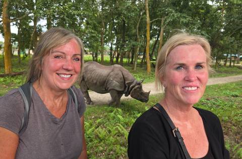Two smiling women stand in front of a grazing one-horned rhinoceros in a lush forest reserve.