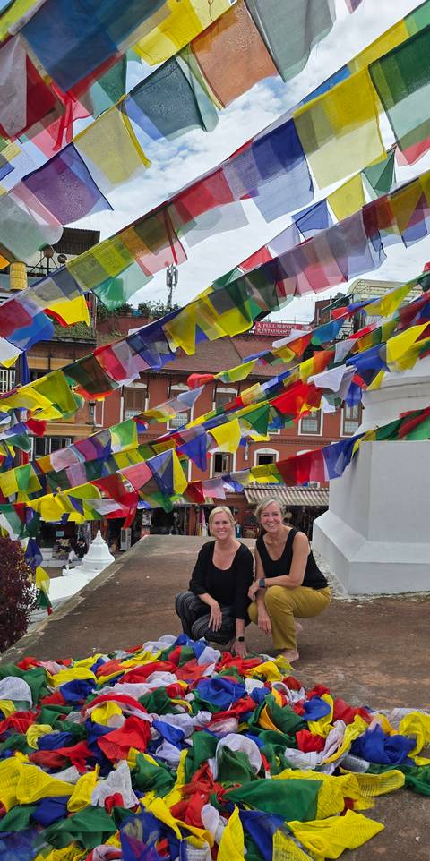 Colorful prayer flags flutter as two women smile beneath them in a vibrant Kathmandu street scene.