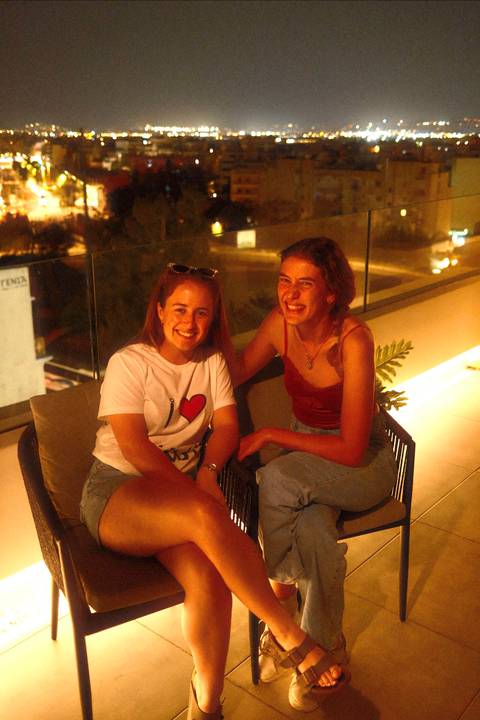 Two young women enjoy drinks together on a softly lit balcony at night.
