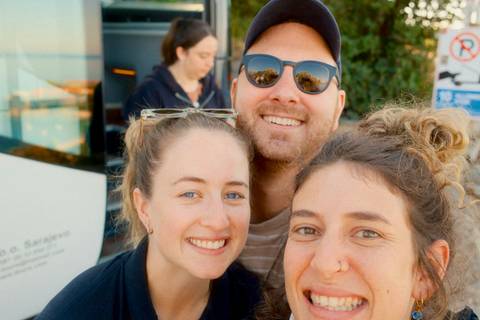 Close-up selfie of three smiling travellers in bright daylight near a tour bus.