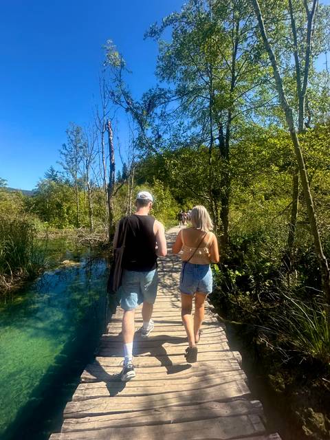 Two travellers walk along a wooden boardwalk over clear water in a lush forest.