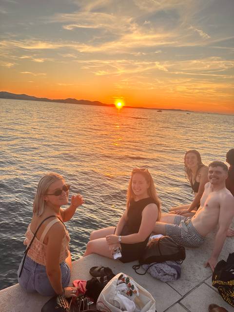 Friends sit on a waterfront platform enjoying a vivid orange sunset over the sea.
