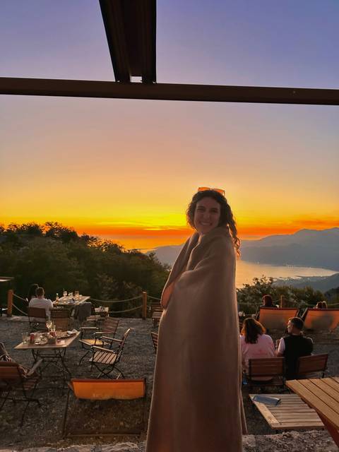 A smiling woman wrapped in a blanket stands against a glowing coastal sunset backdrop.