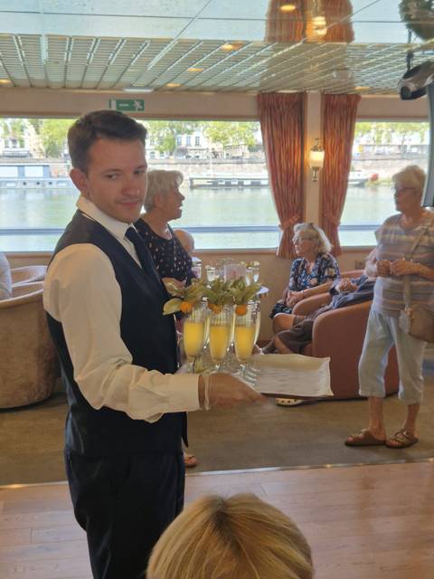 A waiter in formal vest presents champagne flutes to seated passengers inside a river cruise lounge.