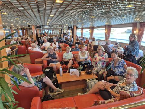 Dozens of cruise passengers relax in a spacious lounge filled with seating and large windows.