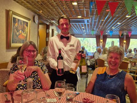 Two guests toast with wine beside a smiling waiter holding multiple bottles in a decorated dining room.
