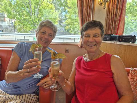 Two women hold up champagne flutes and smile from a comfy river-cruise sofa.