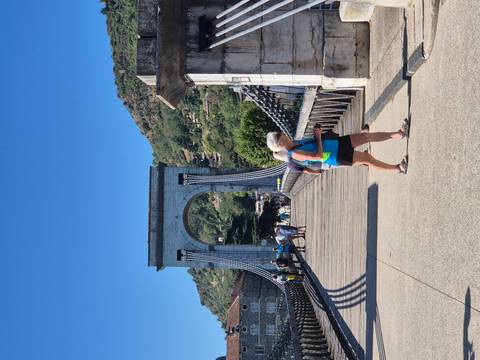 Tourists cross a historic stone-arch suspension bridge on a bright summer day.