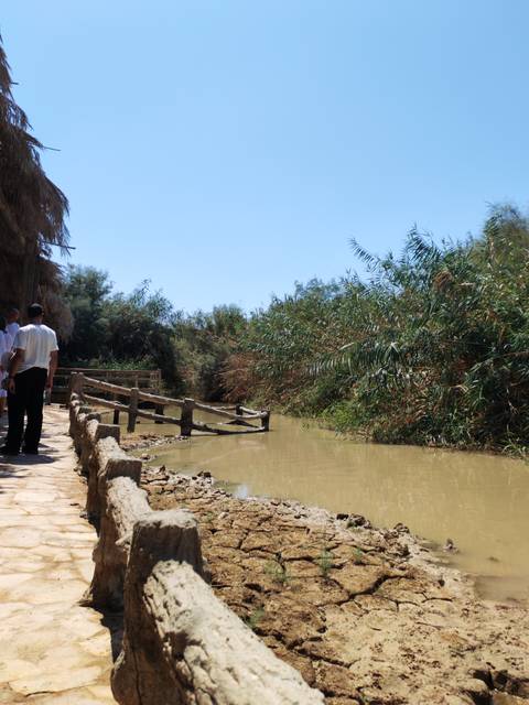 Visitors stand near a muddy river bordered by dense reeds at a historic baptism site.