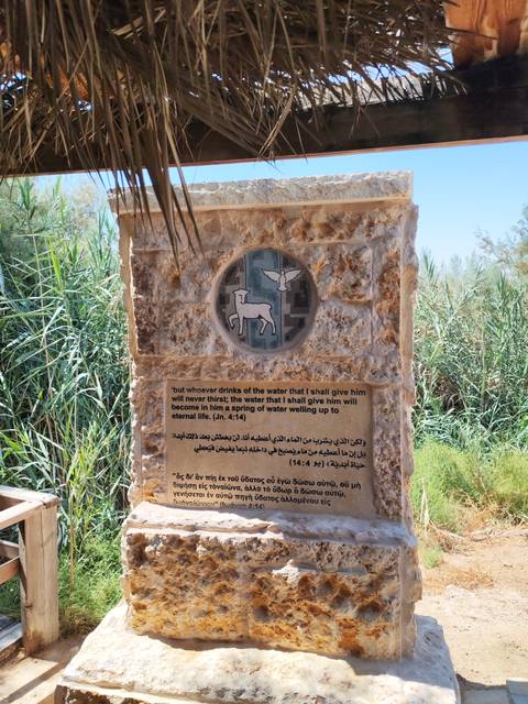 Stone plaque featuring a mosaic of a lamb and dove above a multilingual biblical verse, set outdoors among greenery.
