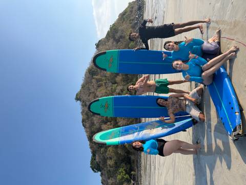 Smiling group posing with blue surfboards on a sunny beach backed by low hills.