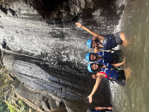 Three adventurers in helmets and life jackets celebrating beneath a cascading jungle waterfall.