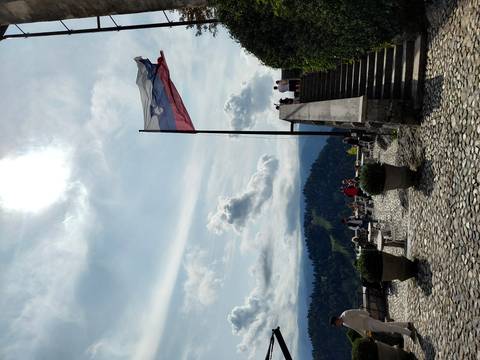 Stone terrace with tables and visitors beneath a flying Slovenian flag, overlooking forested hills and a distant lake.