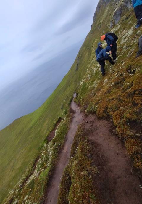 Narrow muddy trail along a steep green cliff above a misty sea, with a hiker ahead.