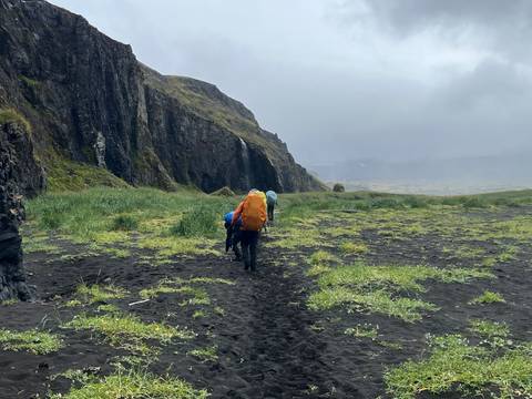 Backpackers trekking across a black-sand valley beneath towering volcanic cliffs on a misty day.