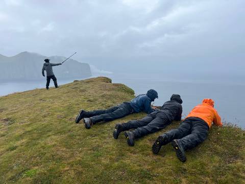 Guide pointing out to sea while companions lie prone at the edge of a grassy cliff in drizzly weather.