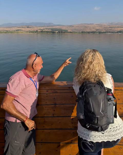 Man points toward open water while talking to a woman with a backpack aboard a wooden boat.