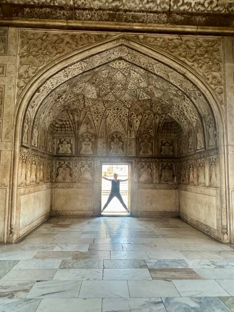 Person poses star-shaped in ornate Mughal interior chamber with intricate stucco ceiling patterns.
