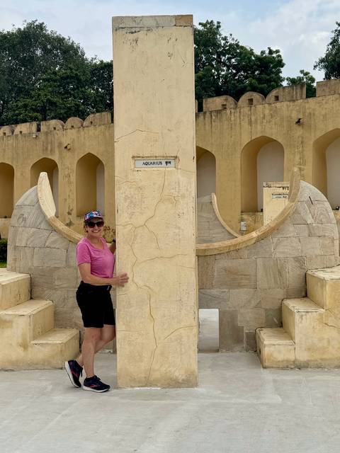 Smiling woman stands beside an engraved zodiac pillar marked 'Aquarius' at Jantar Mantar observatory.