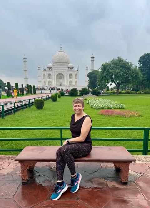 Woman smiles on a bench in lush Mughal gardens with the Taj Mahal rising majestically behind.
