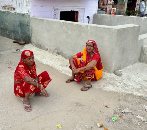 Two local women in bright red saris sit on a roadside against a plain grey wall.