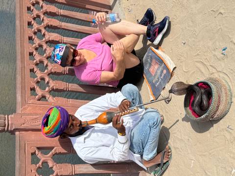 Snake charmer plays a pungi beside a basketed cobra while tourist watches seated on sandy ground.