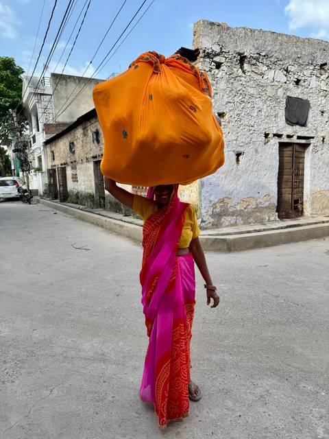 Woman in bright sari walks down rustic street balancing a large orange bundle on her head.