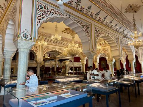 Opulent interior of Jaipur City Palace museum with chandeliers, ornate arches and visitors examining exhibits.