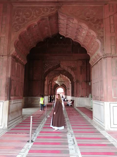 Interior arcade of red sandstone Mughal architecture with hanging chandelier and visitors seated on carpets.