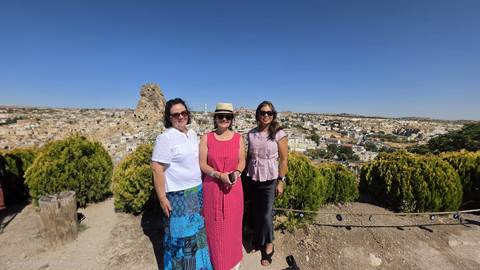 Three women posing at panoramic viewpoint over Cappadocia village and rock formations on clear day.