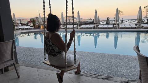 Woman on swing overlooking hotel pool at tranquil sunset with umbrellas and water reflections.
