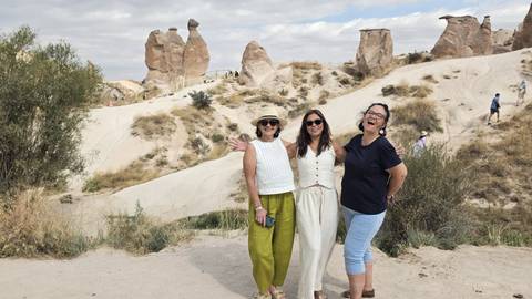 Three women pose smiling in front of Cappadocia’s distinctive fairy-chimney rock formations under a partly cloudy sky.