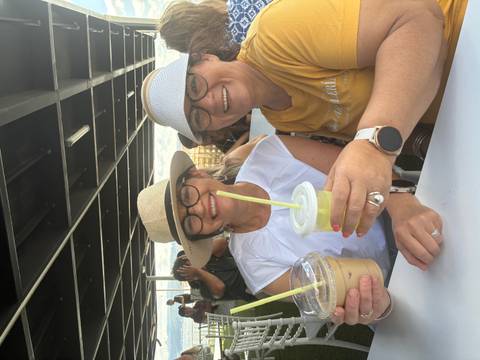 Two women in sunhats toast iced drinks while seated at an outdoor café table.