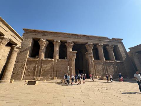 Tourists gather before the grand columned entrance of Edfu Temple under a clear blue sky.