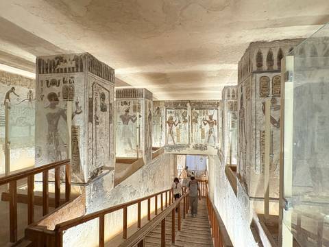 Interior passage of a painted royal tomb with vivid wall reliefs and visitors descending wooden stairs.