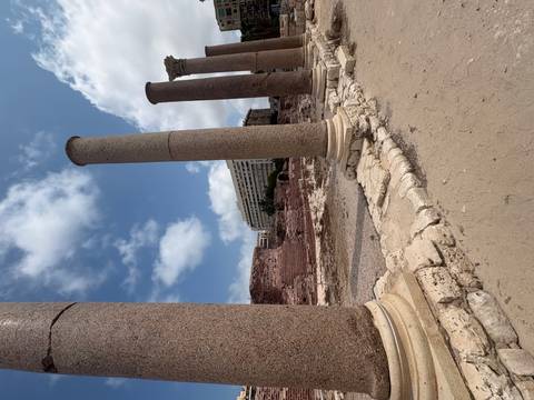 Ruined stone columns frame a modern city building under a partly cloudy sky.