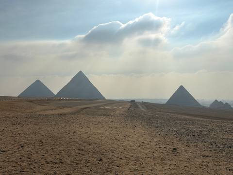 Four pyramids of Giza stand in hazy light as sunbeams filter through clouds above the desert plateau.