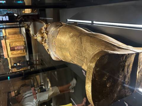 Full-length view of a gilded sarcophagus displayed behind glass in a dim museum hall.