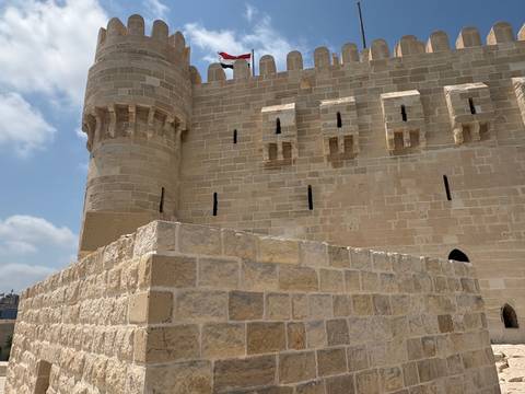 Stone battlements and tower of Qaitbay Citadel flying an Egyptian flag beneath scattered clouds.